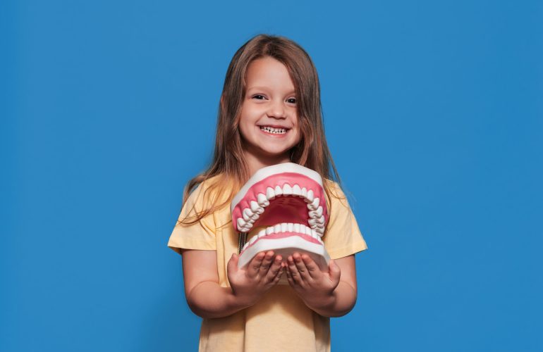A smiling girl with healthy teeth holds a large jaw in her hands on a blue isolated background.Oral hygiene. Pediatric dentistry. Prosthetics. Rules for brushing teeth